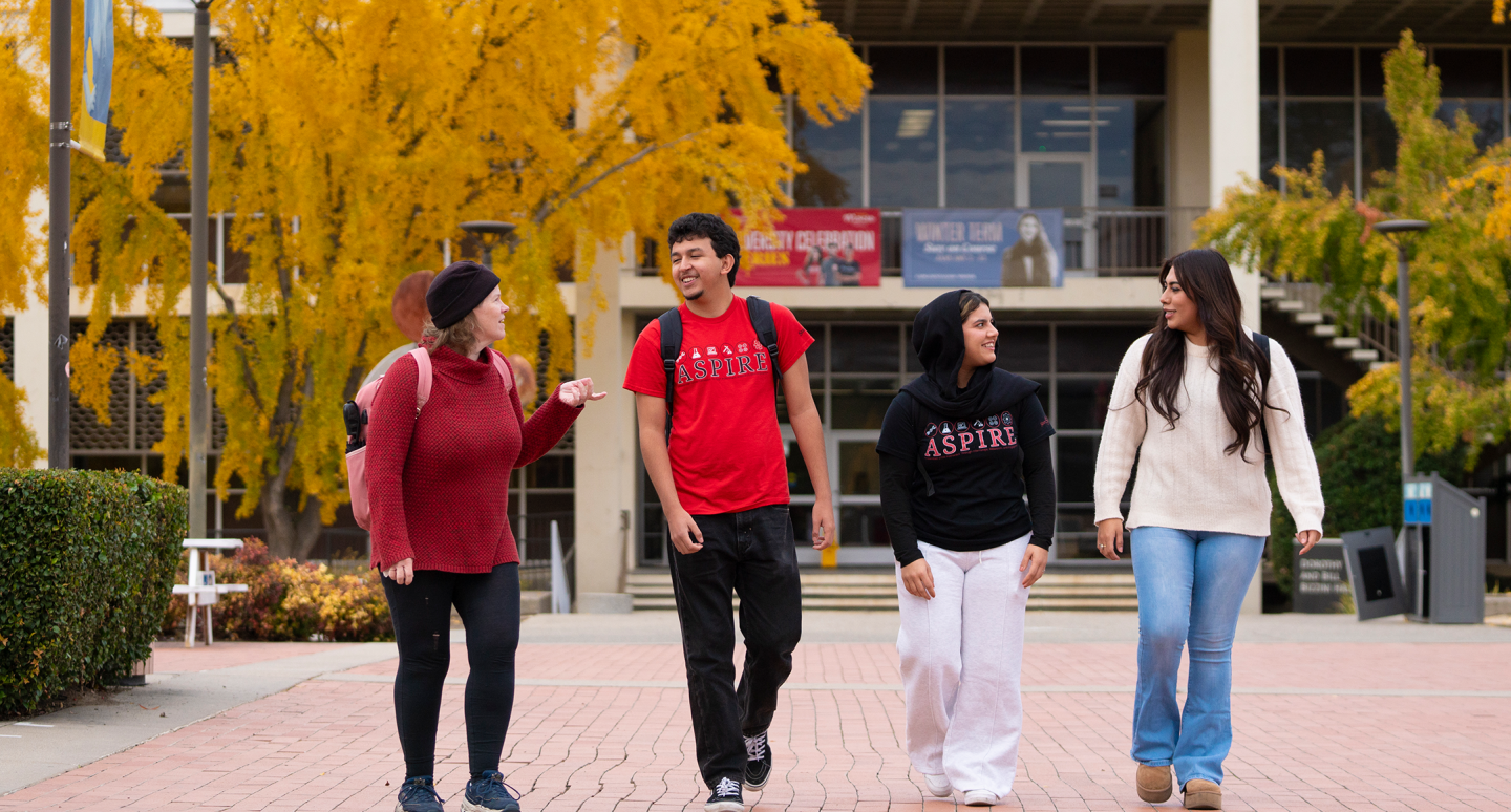 Four people walking and chatting on a campus path lined with yellow-leaved trees, conveying a cheerful and relaxed academic atmosphere.
