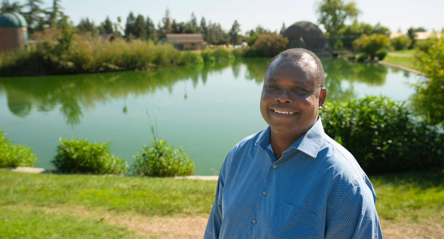 Smiling man in a blue shirt stands by a tranquil pond, surrounded by lush greenery and trees under a clear blue sky, conveying a peaceful vibe.
