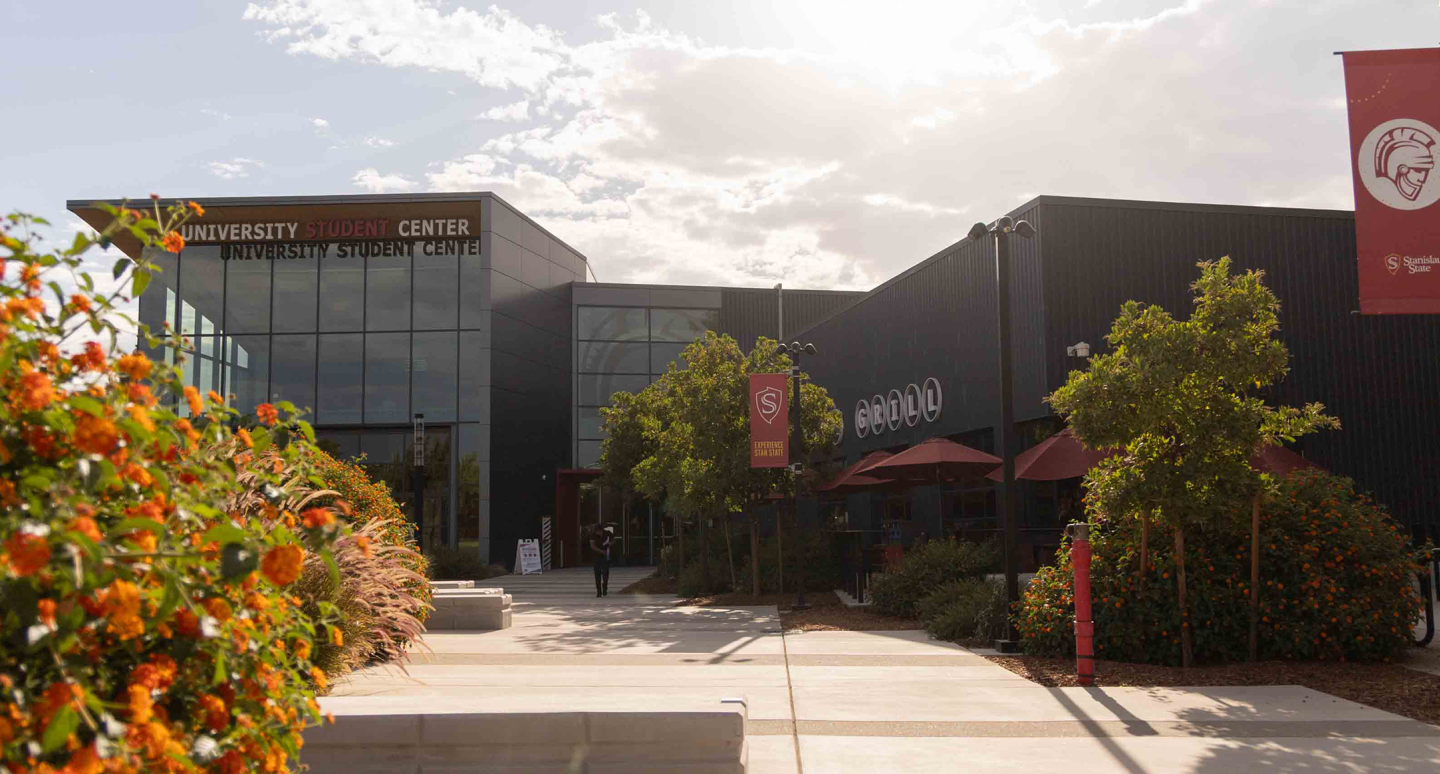 Sunny day outside a modern university student center with glass and dark walls. Orange flowers and green trees line the path leading to the entrance.