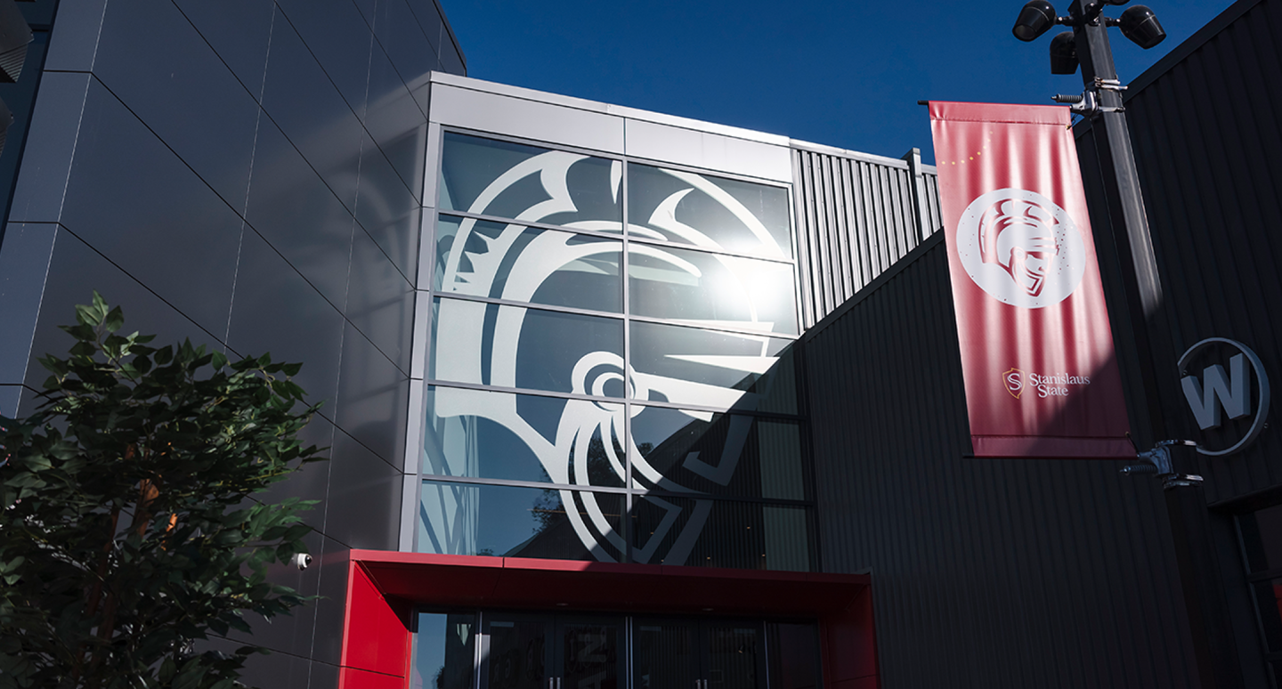Modern building facade with a large stylized graphic of a helmet on glass panels. A red banner waves beside it, under a clear blue sky.