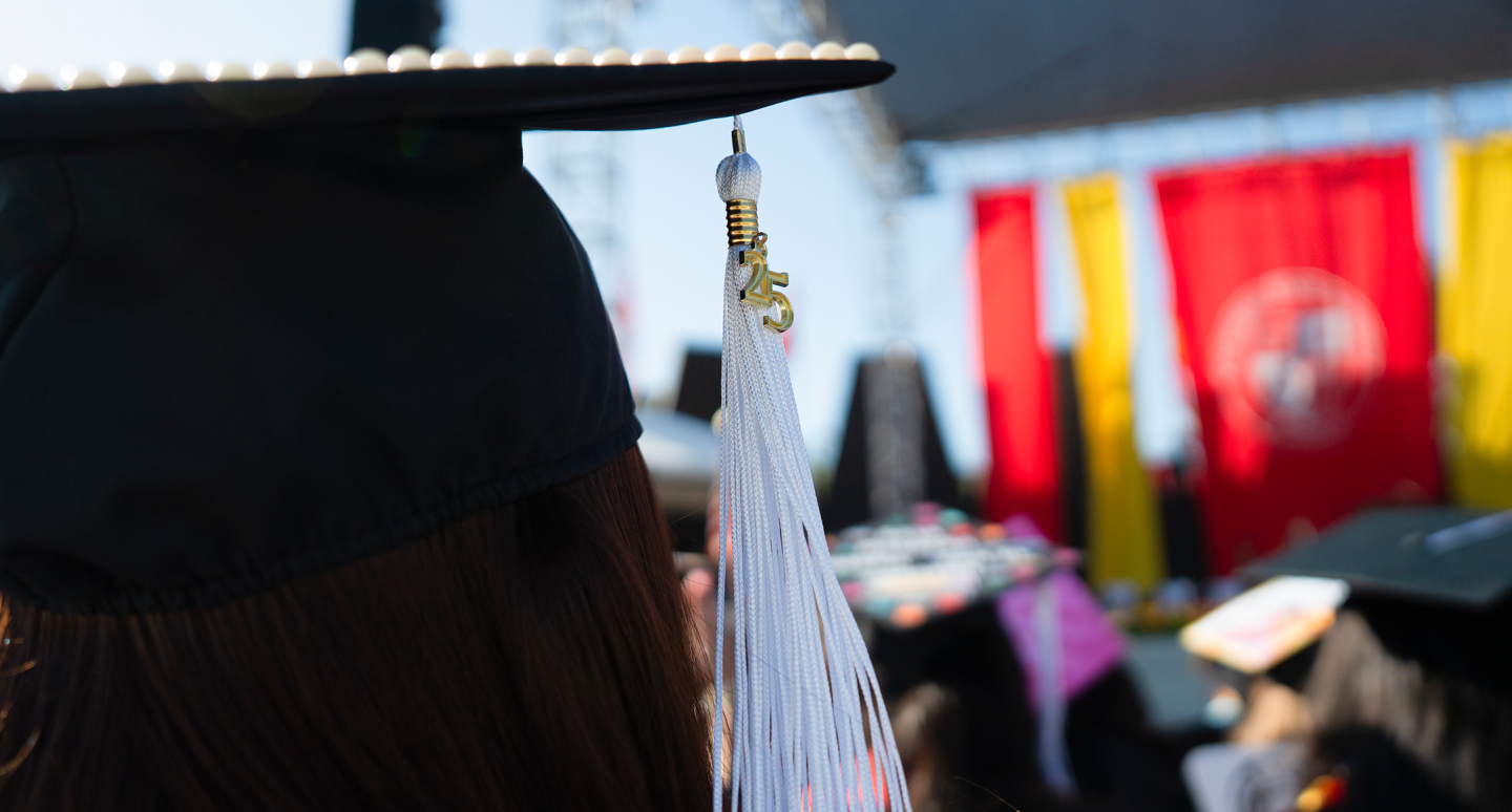 Close-up of a graduate wearing a decorated cap with a white tassel, facing a stage adorned with vibrant banners. The scene conveys achievement and celebration.