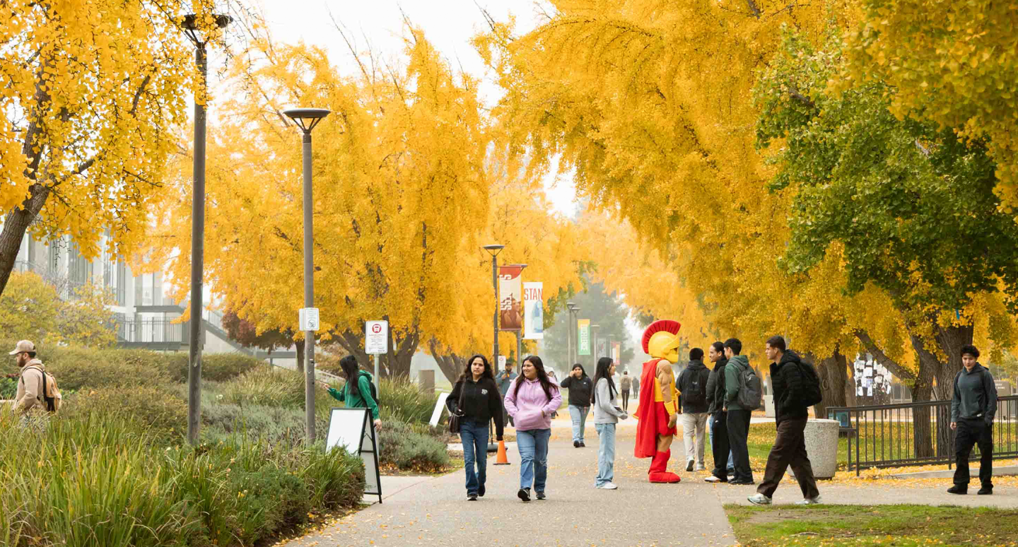 People stroll along a tree-lined path with vibrant yellow foliage. A person in a colorful costume joins them, creating an upbeat and lively atmosphere.