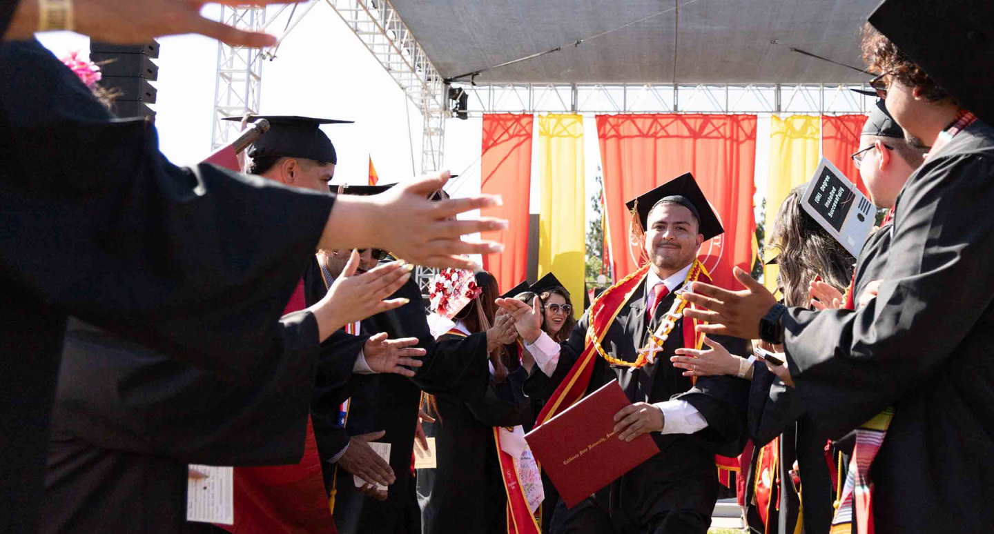 A graduate in cap and gown smiles and holds a diploma, walking through a cheering crowd of peers in similar attire, under a decorated outdoor stage.