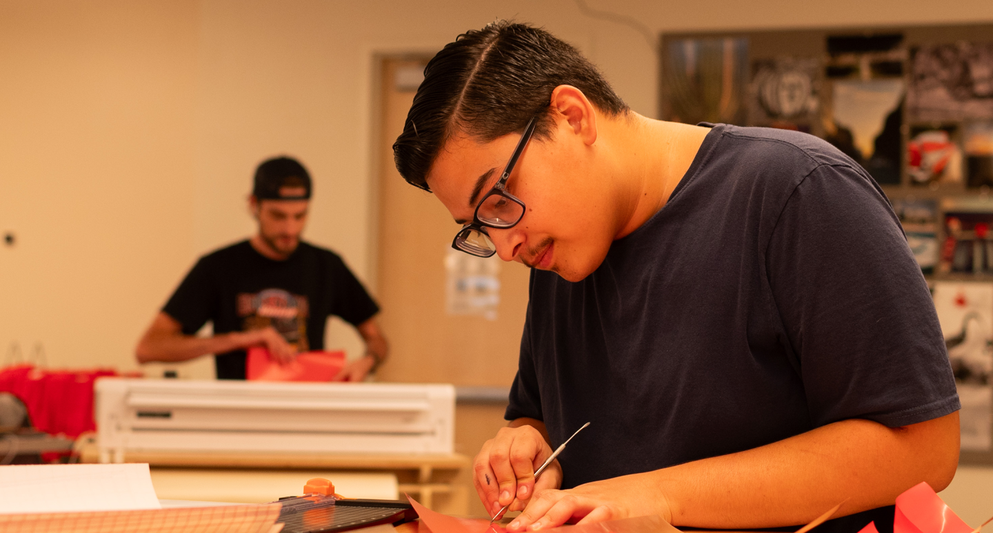 A young man in glasses focuses intently on crafting with paper at a desk. Another person works in the background. The room is warm and well-lit.