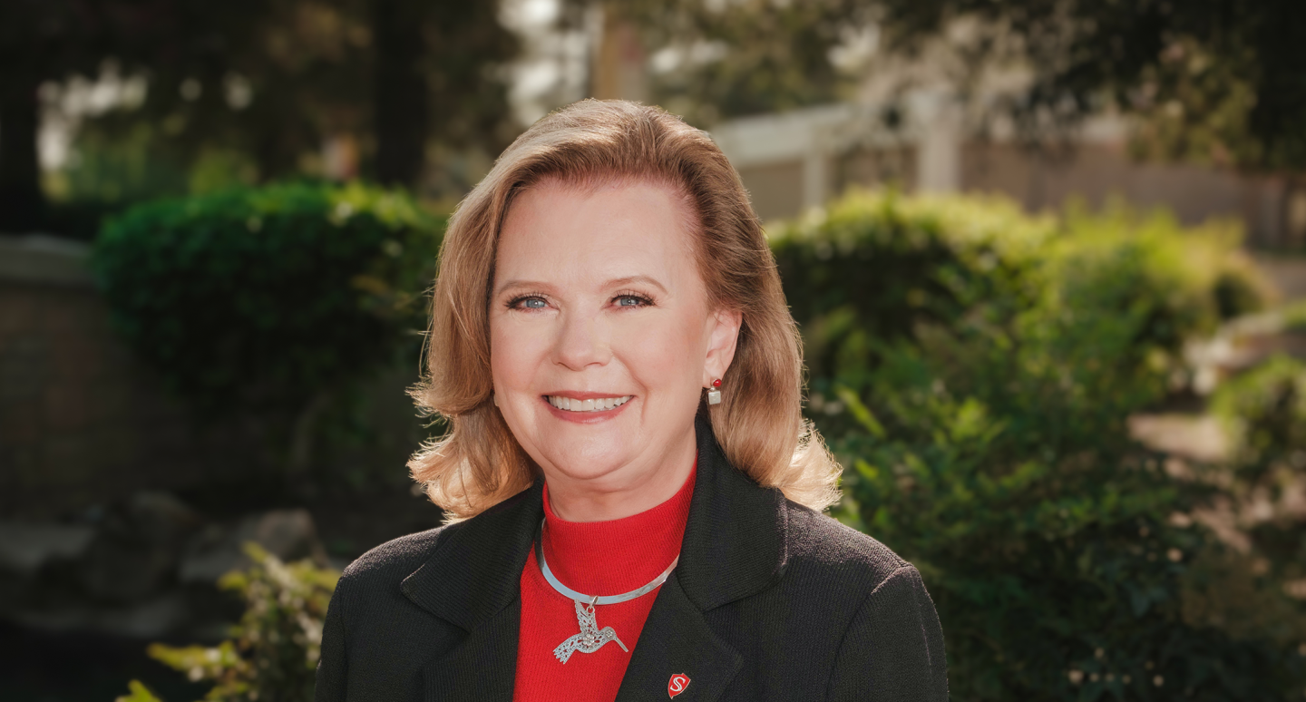 A smiling woman in a black blazer and red top stands outdoors with lush greenery in the background, conveying a warm and professional atmosphere.