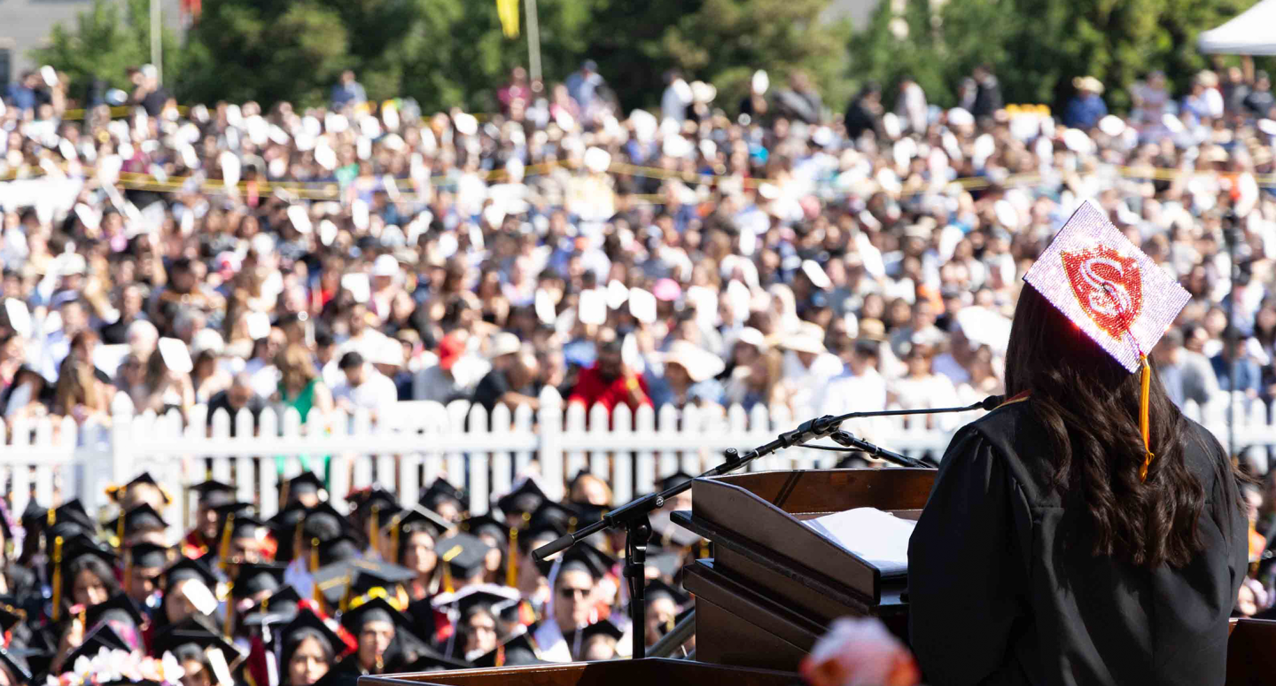 A graduate in a decorated cap speaks at a podium to a large outdoor audience. The crowd, dressed in caps and gowns, fills the sunny space with anticipation.
