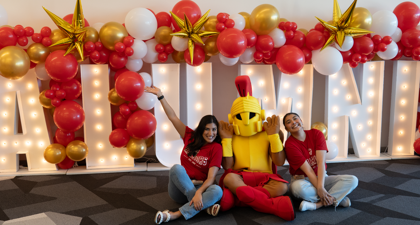 Two people and a large yellow mascot sit cheerfully in front of illuminated "ALUMNI" letters, surrounded by red, gold and white balloons and star decorations.