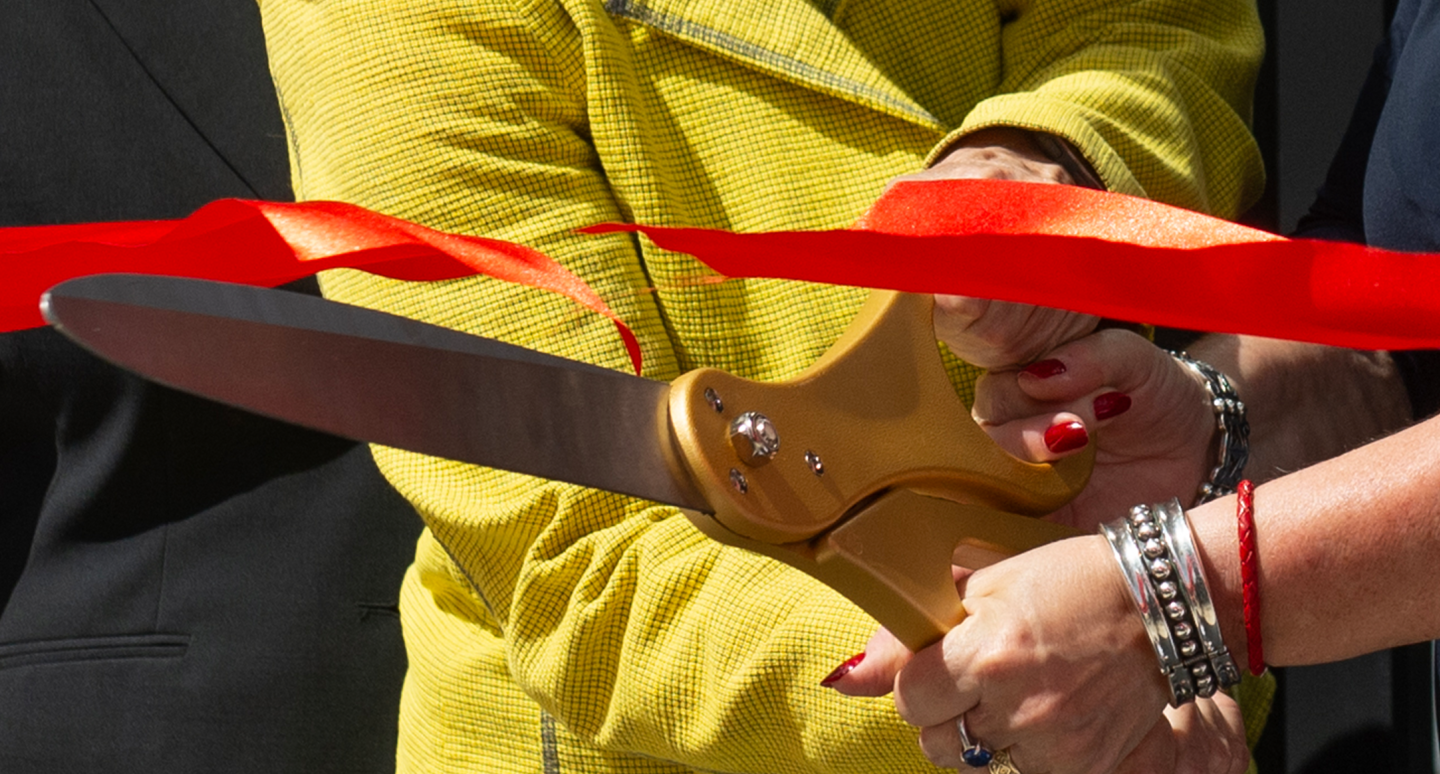 Two people cut a red ribbon with large gold scissors during a ceremony. One wears a yellow jacket, the other's hand has silver bracelets.