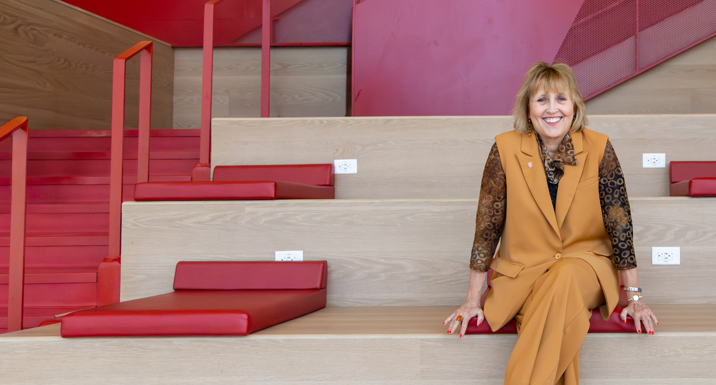 A smiling woman in a mustard-colored suit sits on red-cushioned wooden stairs against a modern, red-accented background. The tone is bright and welcoming.