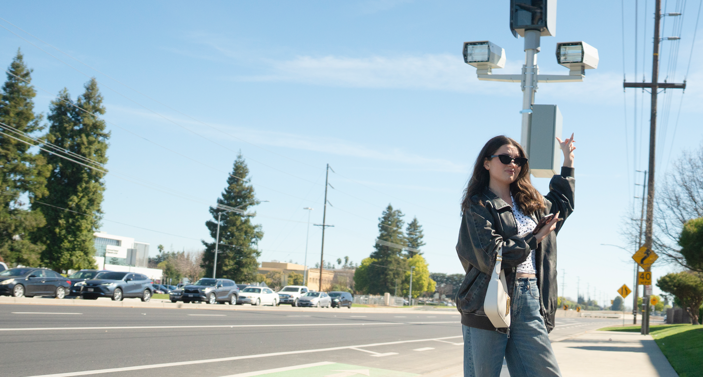 A woman wearing sunglasses stands on a sunny roadside, gesturing towards a traffic light. Cars and trees line the street, conveying a casual, lively urban scene.