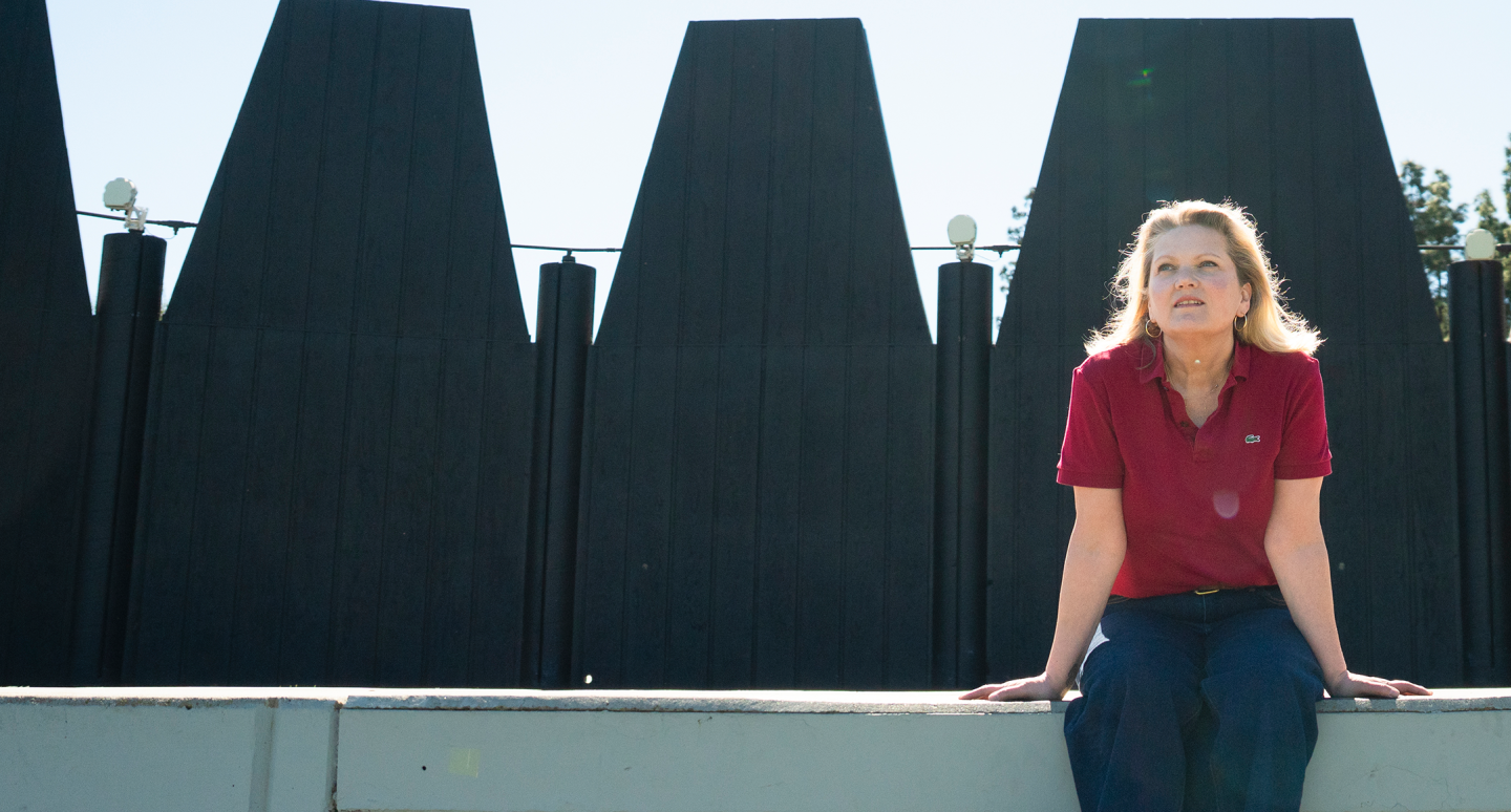 A woman in a red shirt and jeans sits on a concrete ledge against a backdrop of tall, dark geometric sculptures. She gazes upward with a serene expression.
