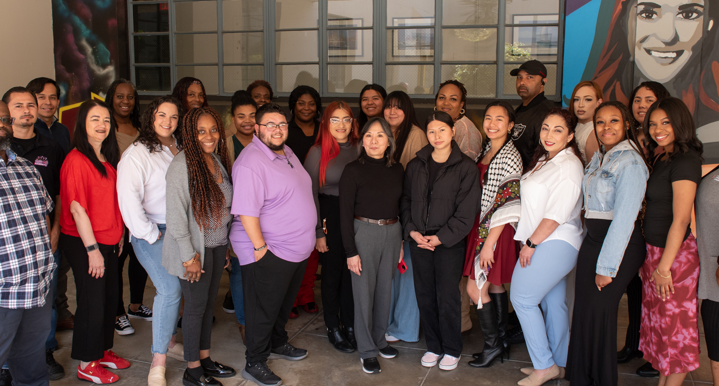Diverse group of smiling colleagues pose indoors in front of colorful murals and tall windows, creating a warm, celebratory atmosphere.