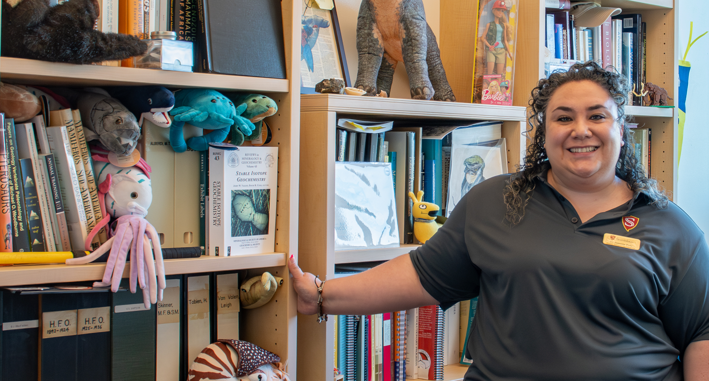 Smiling woman in a black polo stands beside packed bookshelves filled with books, binders, and stuffed animals in a bright library.