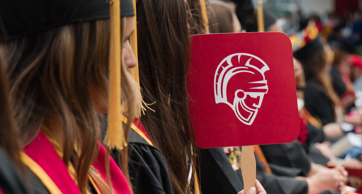 Graduates in black caps and gowns sit outdoors, holding a red sign with a white Spartan helmet emblem, creating a proud, celebratory mood.