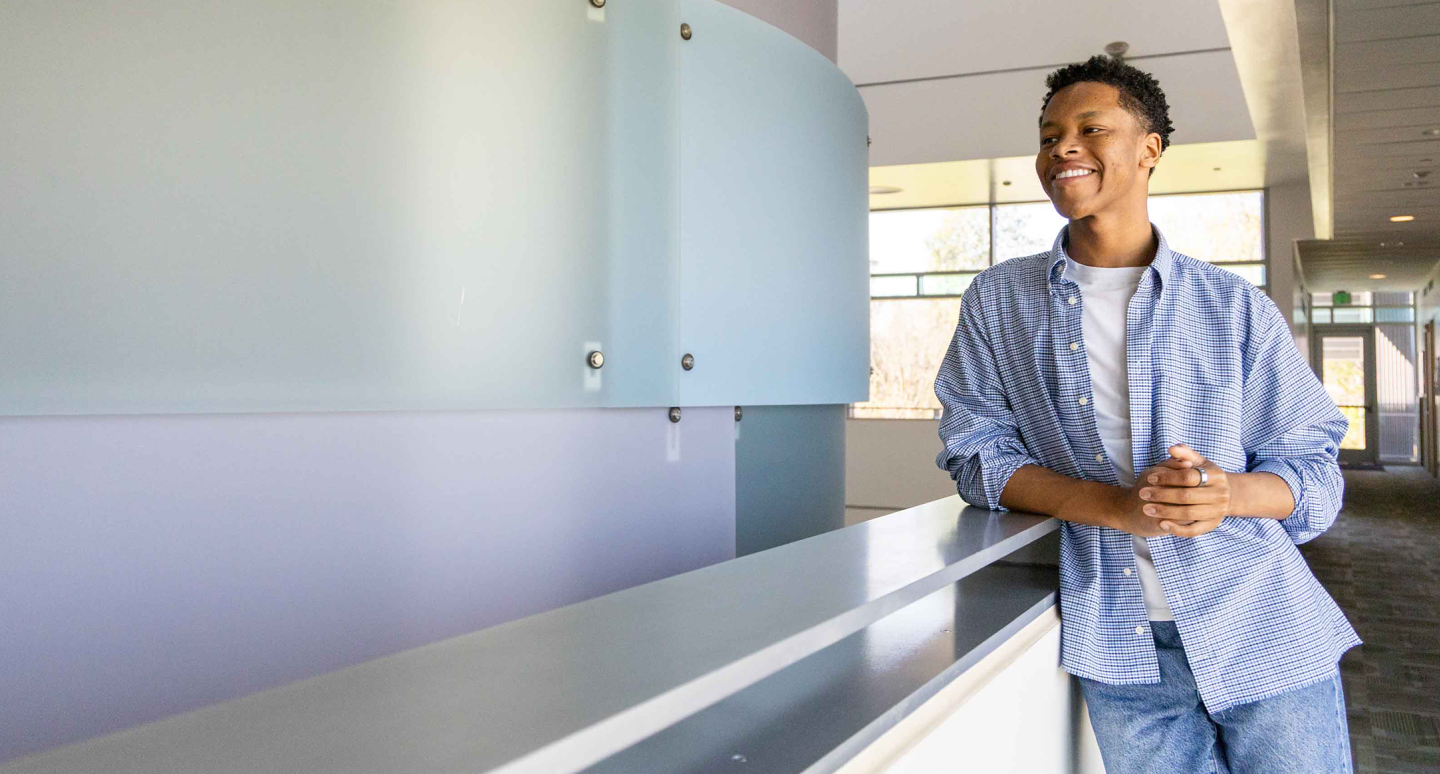 Smiling person in casual attire leans on a white railing in a bright, modern hallway. Sunlight streams through large windows, creating a cheerful ambiance.
