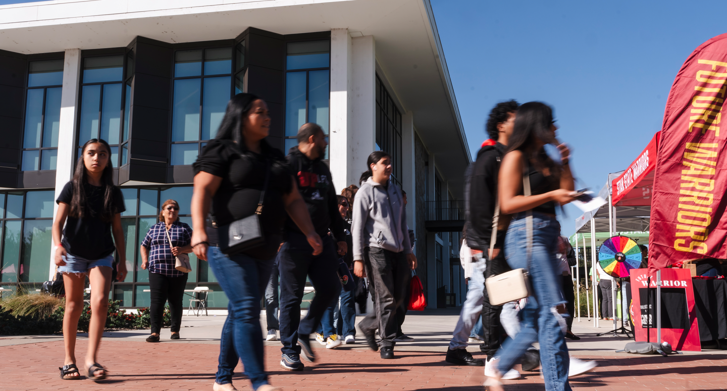Students walk past a modern campus building toward colorful event booths and banners in bright sunshine, creating a lively, welcoming atmosphere.
