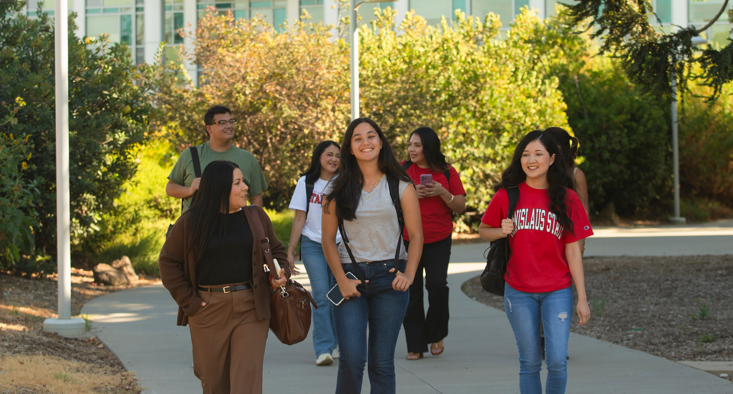 A diverse group of six students walks along a sunny campus path, chatting and smiling amid leafy trees and modern buildings; the scene feels relaxed and welcoming.