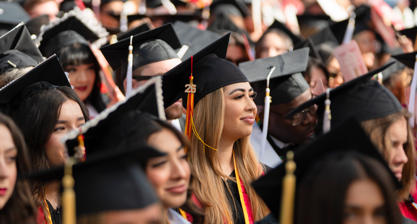  Graduates in black caps and gowns gather closely, smiling and celebrating at a lively commencement ceremony with colorful decorations.