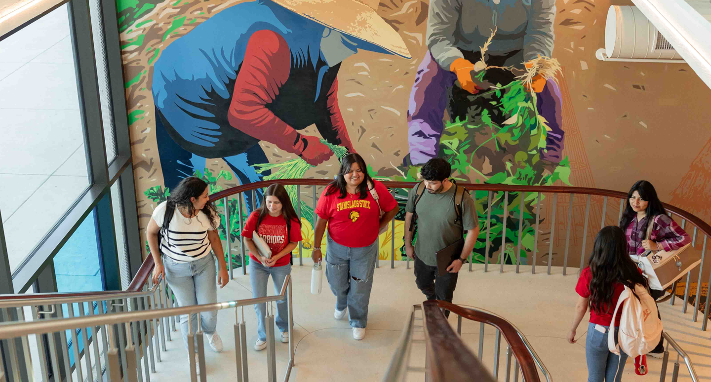 Students walk up a school stairway beside a vibrant mural of dancers, with metal railings, large windows, and a lively communal atmosphere.