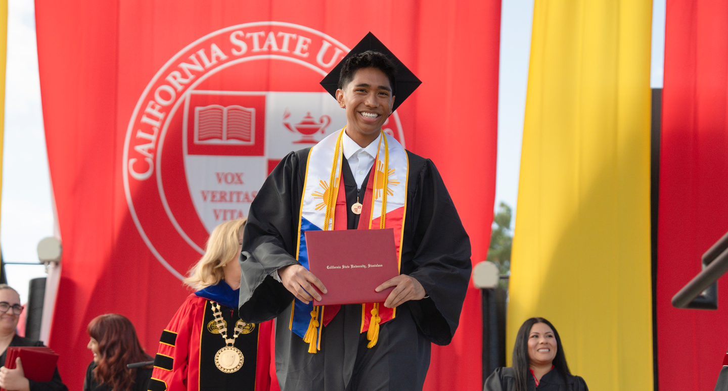 A smiling graduate in cap and gown holds a diploma before a California State University seal, with classmates and faculty nearby at commencement.