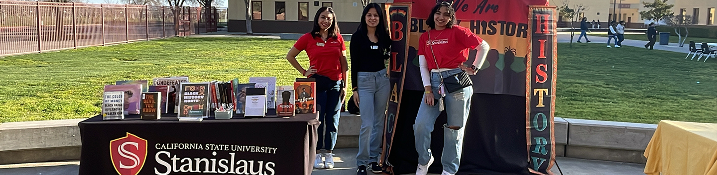 Three students stand near a table holding books.