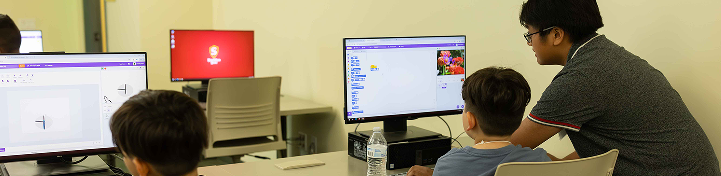 Two young students are seated at separate computers while an instructor offers guidance to one of them.