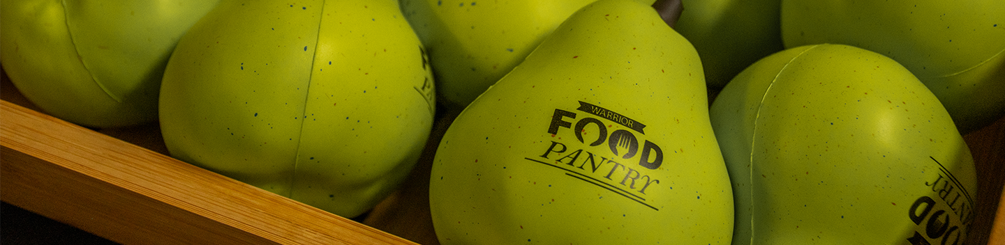 Close-up of green pear-shaped stress balls with "Warrior Food Pantry" text, nestled in a wooden crate. Bright, playful, and community-focused tone.
