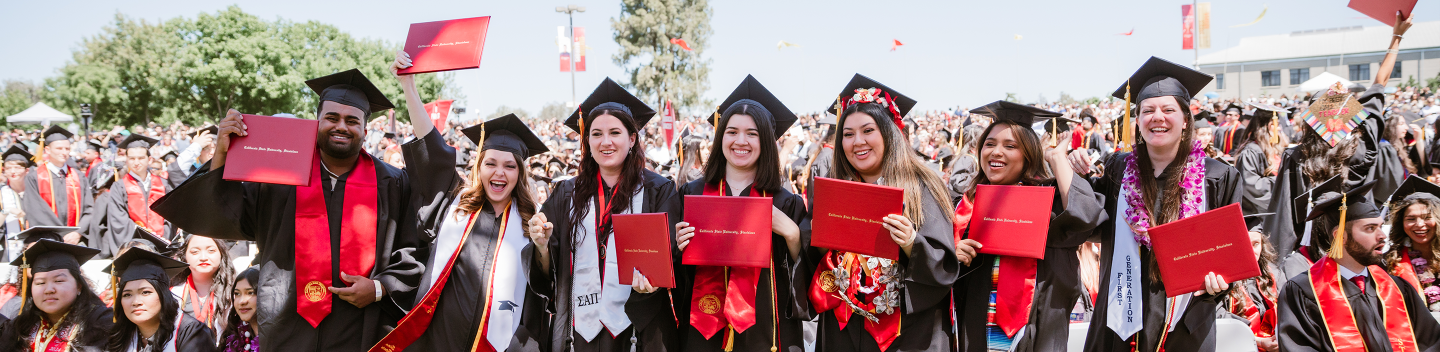 A group of seven graduates in caps and gowns joyfully hold diplomas, celebrating on a sunny day outdoors, surrounded by a large, cheering crowd.