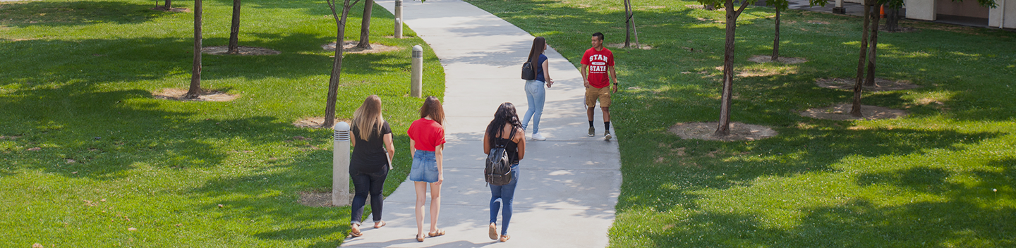 A group of four students walks on a tree-lined campus path, while another student approaches wearing a red shirt. The scene conveys a relaxed, sunny atmosphere.