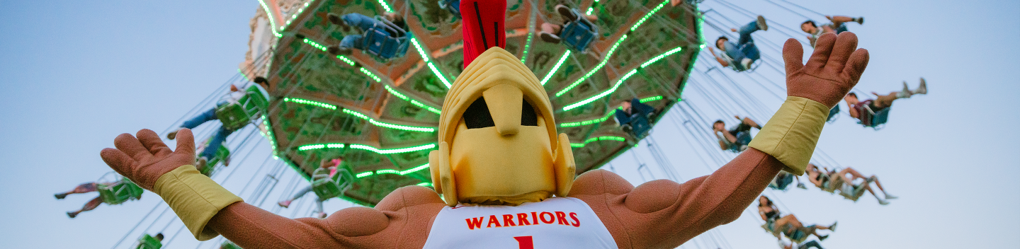 Titus stands with arms wide open in front of a colorful swing ride. People are joyfully swinging high in the air.