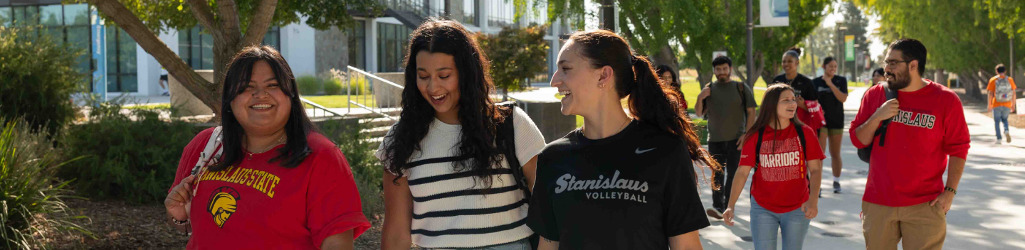 Students walking together on a sunny Stanislaus State campus pathway, smiling and talking as they head to class, with others walking in the background.