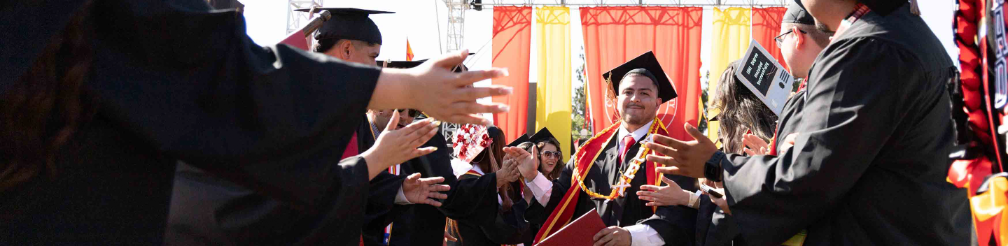 A graduate in cap and gown smiles and holds a diploma, walking through a cheering crowd of peers in similar attire, under a decorated outdoor stage.