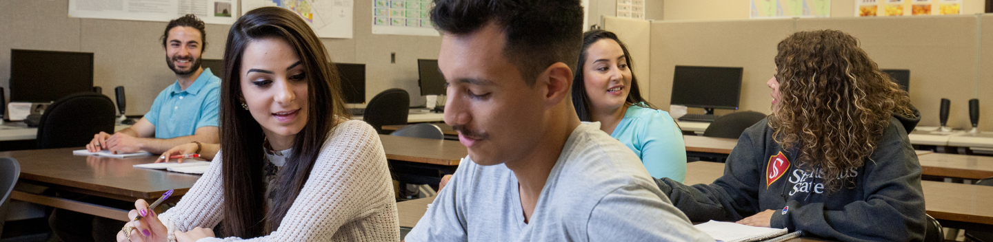 A diverse group of students sits in a classroom, engaged in discussion. Papers and computers are on the desks, creating a collaborative and focused atmosphere.
