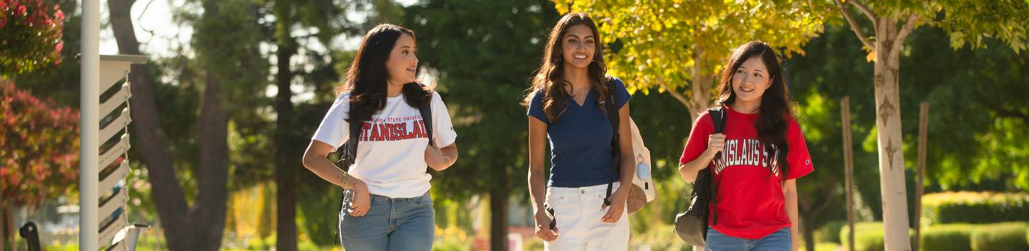 Three Stanislaus State students walk together across campus on a sunny day, carrying backpacks and wearing University apparel.