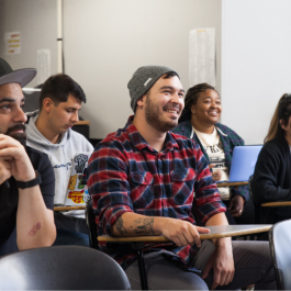 College students sitting in a classroom, listening and smiling during a lesson, with desks, notebooks, and a laptop visible.