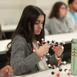 A college student sitting in a classroom, studying a molecular model during a hands-on lesson, with other students listening in the background