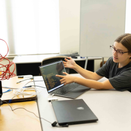 A student in a computer lab examining code on a laptop and interacting with networking equipment during a hands-on tech project.