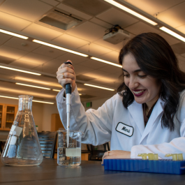 A student in a chemistry lab wearing a lab coat and using a pipette to transfer liquid into a beaker, with glassware and lab equipment on the table.