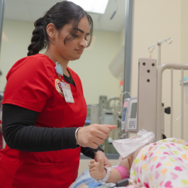 A Stanislaus State nursing student in red scrubs practices patient care in a clinical lab with a pediatric mannequin.