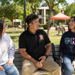 Three Stanislaus State students sit together on a bench in the campus quad, smiling and talking on a sunny day.