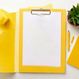 A yellow clipboard with a blank paper, next to a small plant and notebook