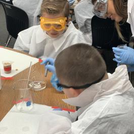 Students wearing safety goggles, gloves, and lab coats conduct a hands-on science experiment at a classroom table while an instructor leans in to guide them.