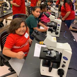 Students sit at lab tables using microscopes in a science classroom while instructors in red shirts assist in the background.