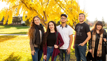 Five students pose for a group photo beside beautiful autumn trees.