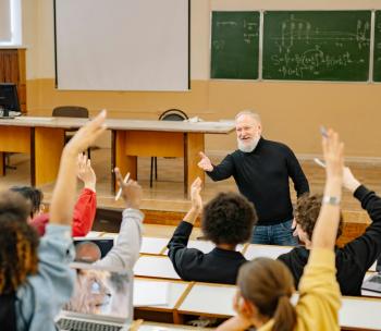 Picture of a faculty member in class talking with his students. The students have their hands raised as if in response to what he's saying. 