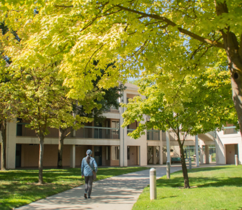 a female student walks on a pathway through trees, with a three story residential in the background 