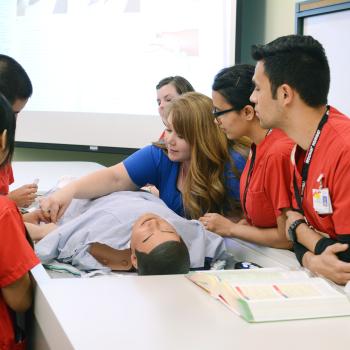 Medical students in red scrubs gather around a mannequin for a hands-on lesson, while an instructor in blue demonstrates techniques. The setting is educational.