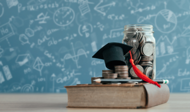 a book with change, pencils and a graduation cap stacked on top. With a chalkboard in the background.