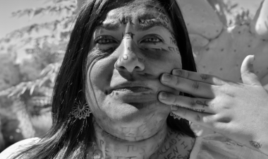 Black and white photograph depicting a woman focused on the camera. It's titled Calladita Te Ves Más Bonita by Artist Monica Crystal Ocegueda.