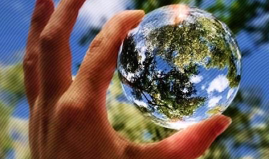 Hand holding a clear, glass ball with blue sky and trees in the background.